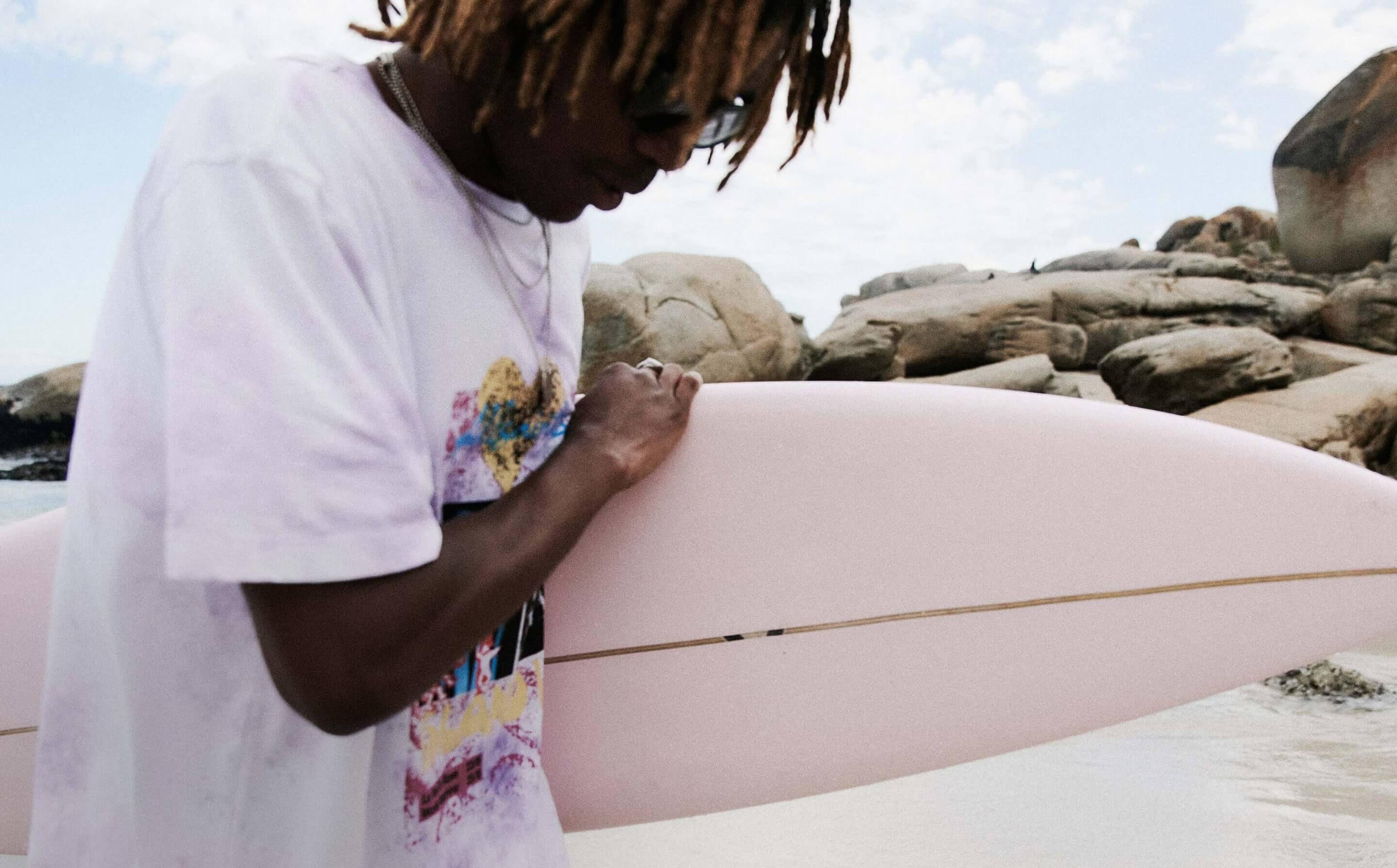 A person with dreadlocks wearing a white graphic T-shirt and sunglasses holds a pale pink surfboard near rocky shore and ocean water.