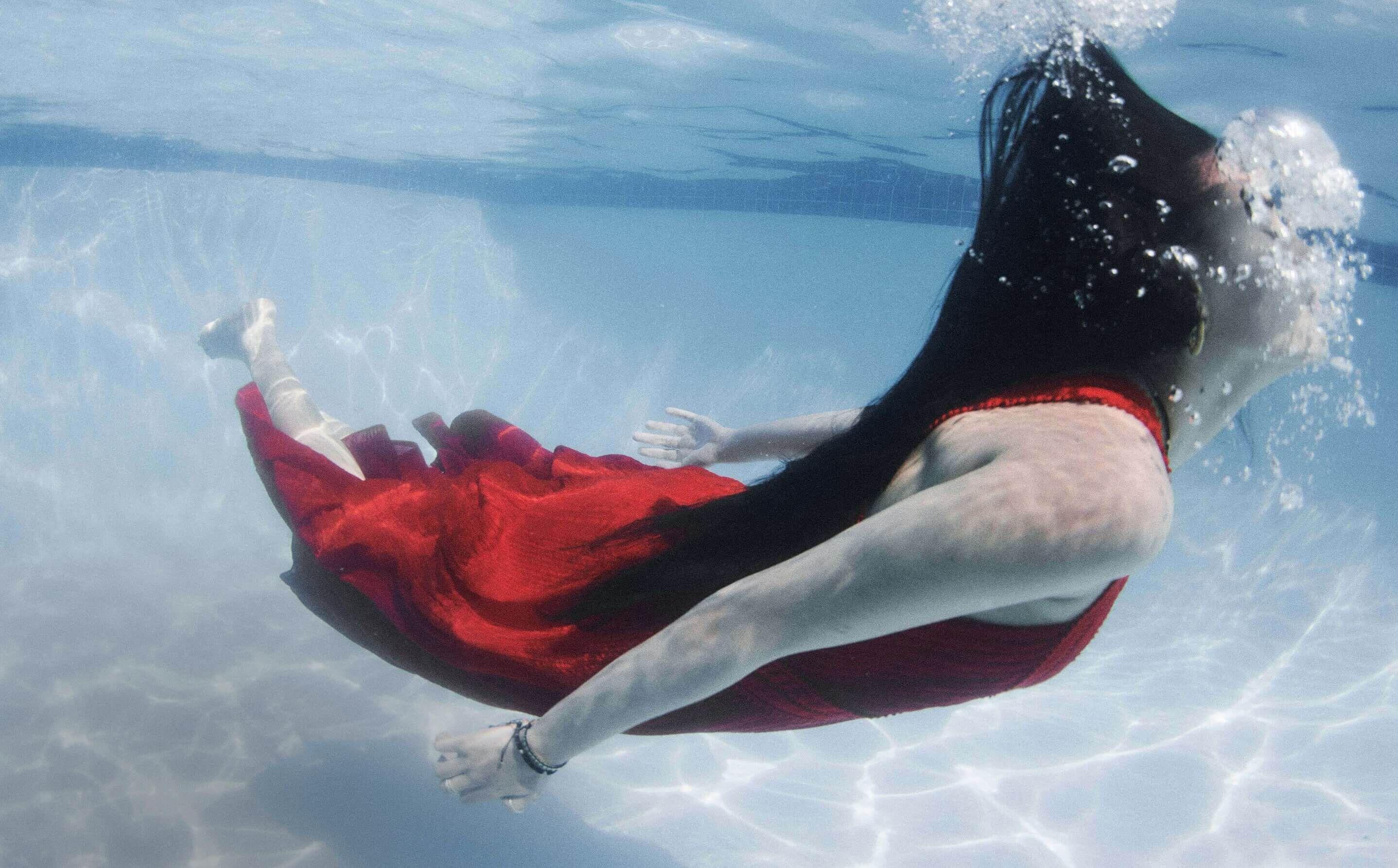 Underwater shot of a person swimming horizontally. They have long black hair flowing behind them and are wearing a flowing red dress. Bubbles are visible around their head, and the pool floor and walls are faintly visible through the clear water.