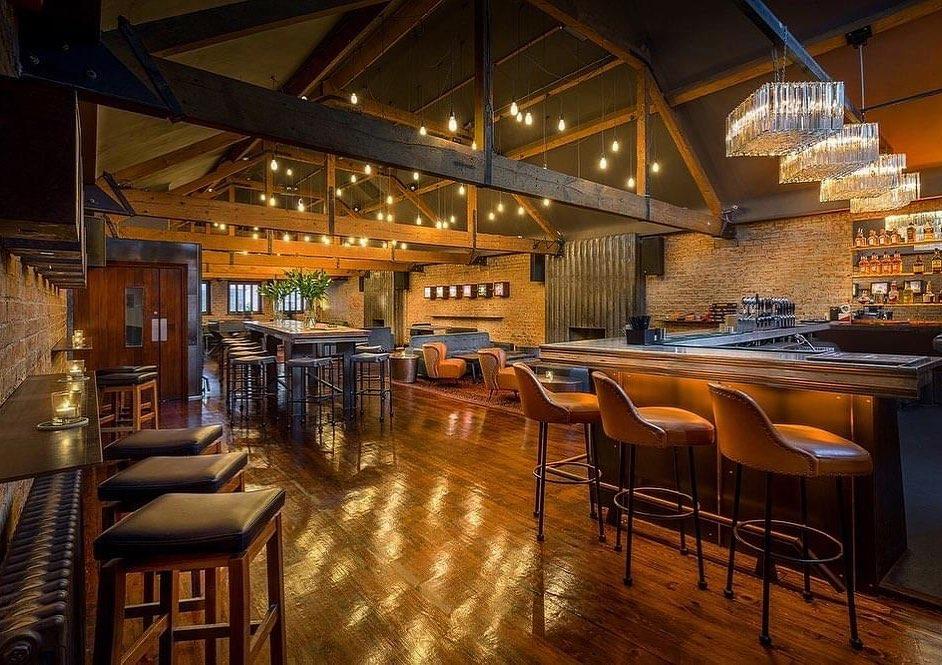 A warm, inviting bar interior featuring polished wooden floors and exposed wooden beams on the ceiling. The bar counter has tan cushioned stools, and there are additional seating areas with tables and chairs. Soft hanging lights illuminate the space, highlighting the exposed brick walls and a display of bottles behind the bar. The overall ambiance is cozy and elegant.
