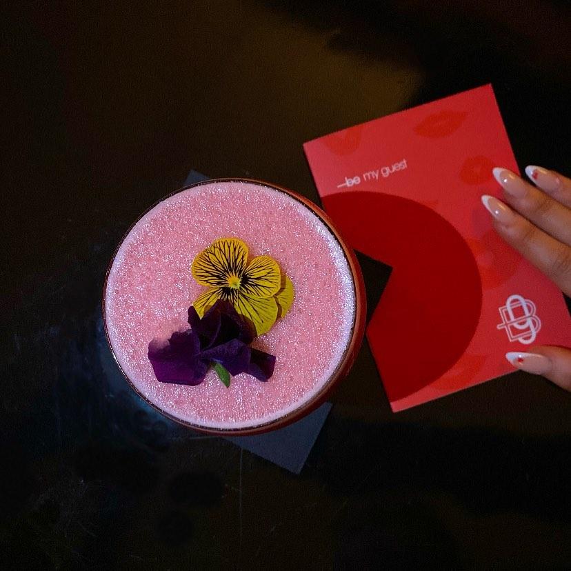 Overhead view of a pink frothy cocktail garnished with a yellow and a purple edible flower, set on a black surface. To the right, a hand with manicured nails holds a red card that reads "—bee my guest" with a white bee logo.