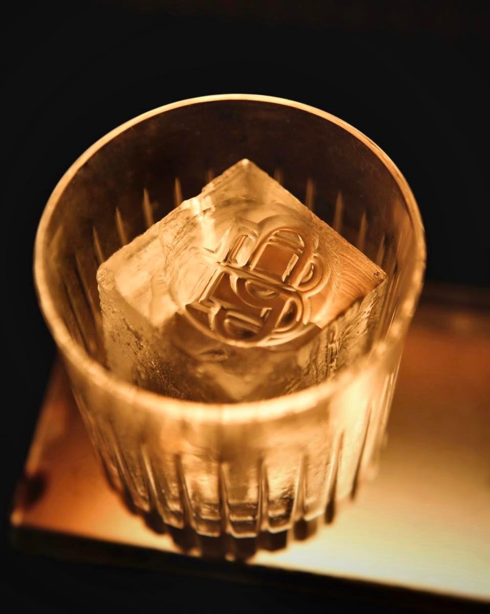 A close-up of a glass with vertical ridges containing a single large ice cube. The ice cube has an intricate design or logo engraved on its top surface. The glass and ice are illuminated with warm, golden lighting against a dark background.