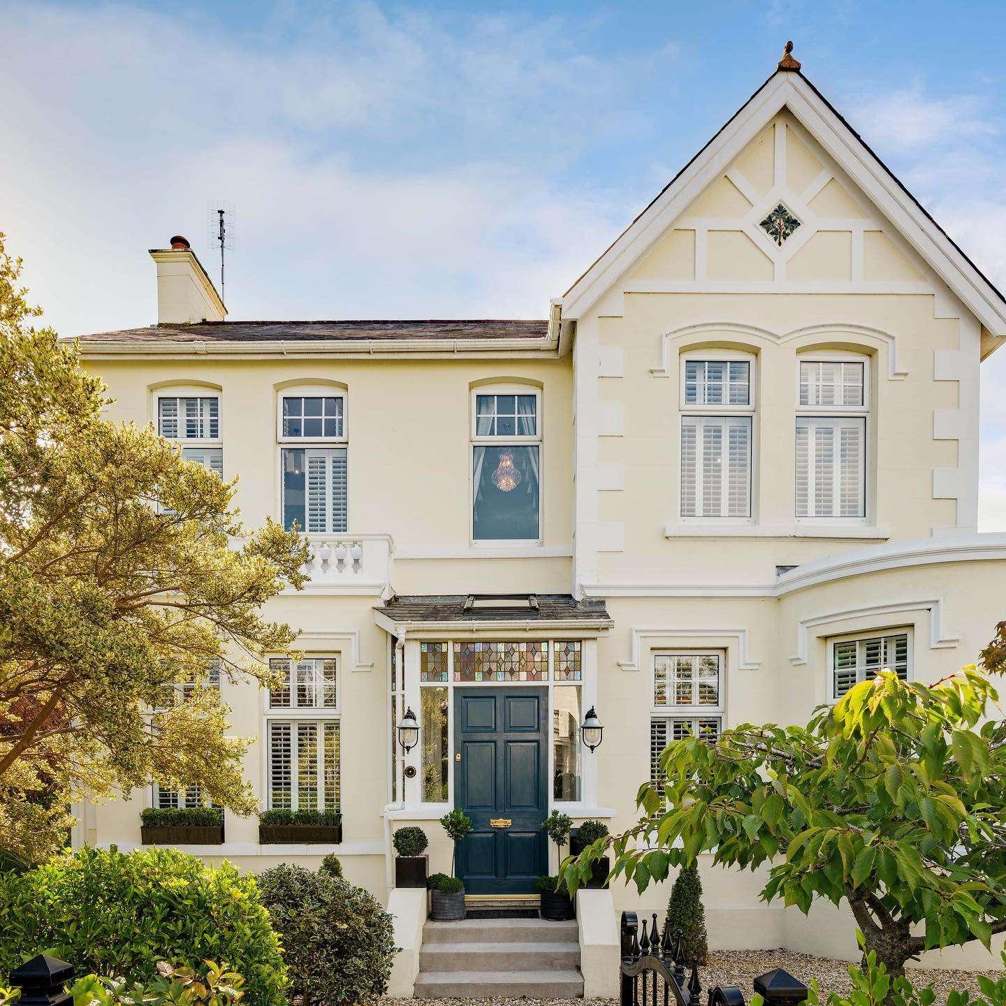 Front view of a large cream-colored Victorian-style house with white trim, a dark green front door, and symmetrical windows with white shutters. The entrance is framed by stained glass panels and black lanterns, surrounded by well-manicured greenery and a small garden path.