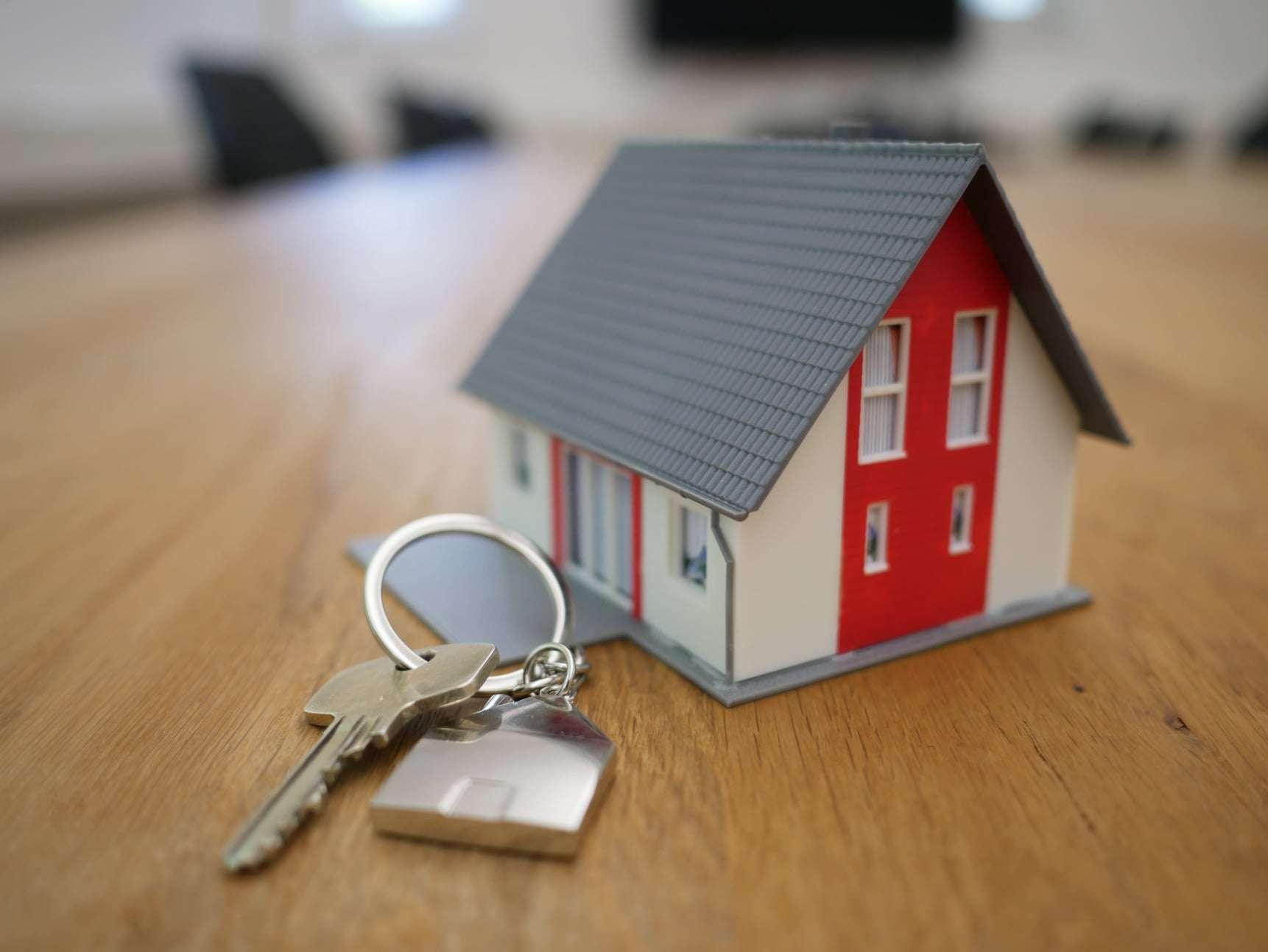Close-up of a small model house with red and white walls and a gray roof, placed on a wooden surface. A silver house key attached to a house-shaped keychain is lying next to the model, symbolizing real estate or home ownership. The background is blurred, suggesting an indoor office or meeting room setting.