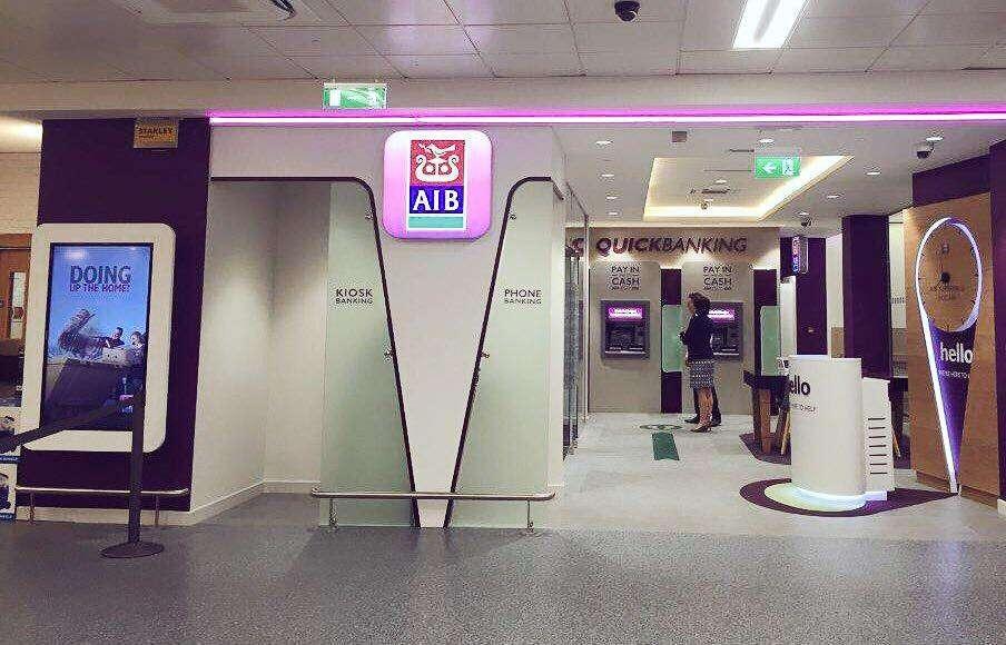 Interior view of an AIB (Allied Irish Banks) branch. The area includes self-service kiosks, a phone banking booth, and multiple ATMs under a "Quickbanking" sign. A woman is using one of the ATMs. The space is brightly lit with purple accent lighting and modern signage. A digital advertisement screen is visible on the left.