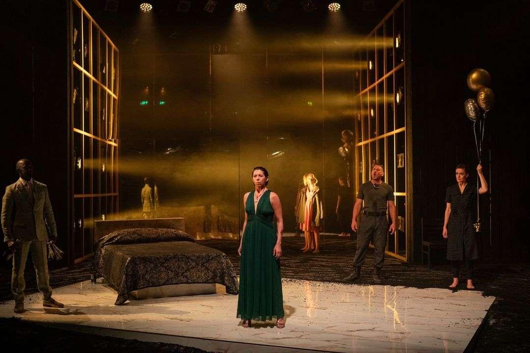 Five actors on stage in a dramatic scene. A woman in a green dress stands center front on a white floor next to a bed with black and gold bedding. To her left, a man in a light suit holds papers. Behind her, a woman in a coat stands near a door with bright light. To her right, a man in dark clothes appears to be shouting, and a woman in black holds gold balloons. The stage has tall panels and warm, focused lighting.