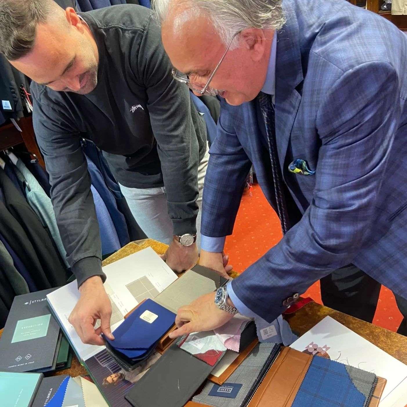 Two men looking at a variety of fabric swatches on a table inside a tailor shop, one wearing a casual outfit and the other in a blue checked suit, both discussing material options.