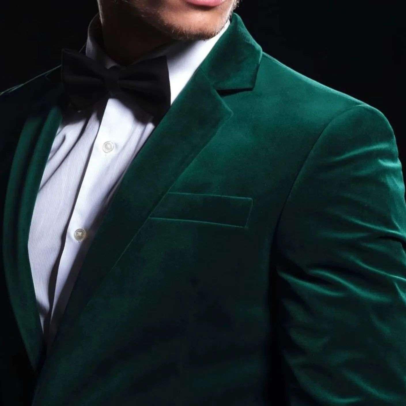 Close-up of a man wearing a deep green velvet tuxedo jacket with a white dress shirt and black bow tie, set against a dark background.