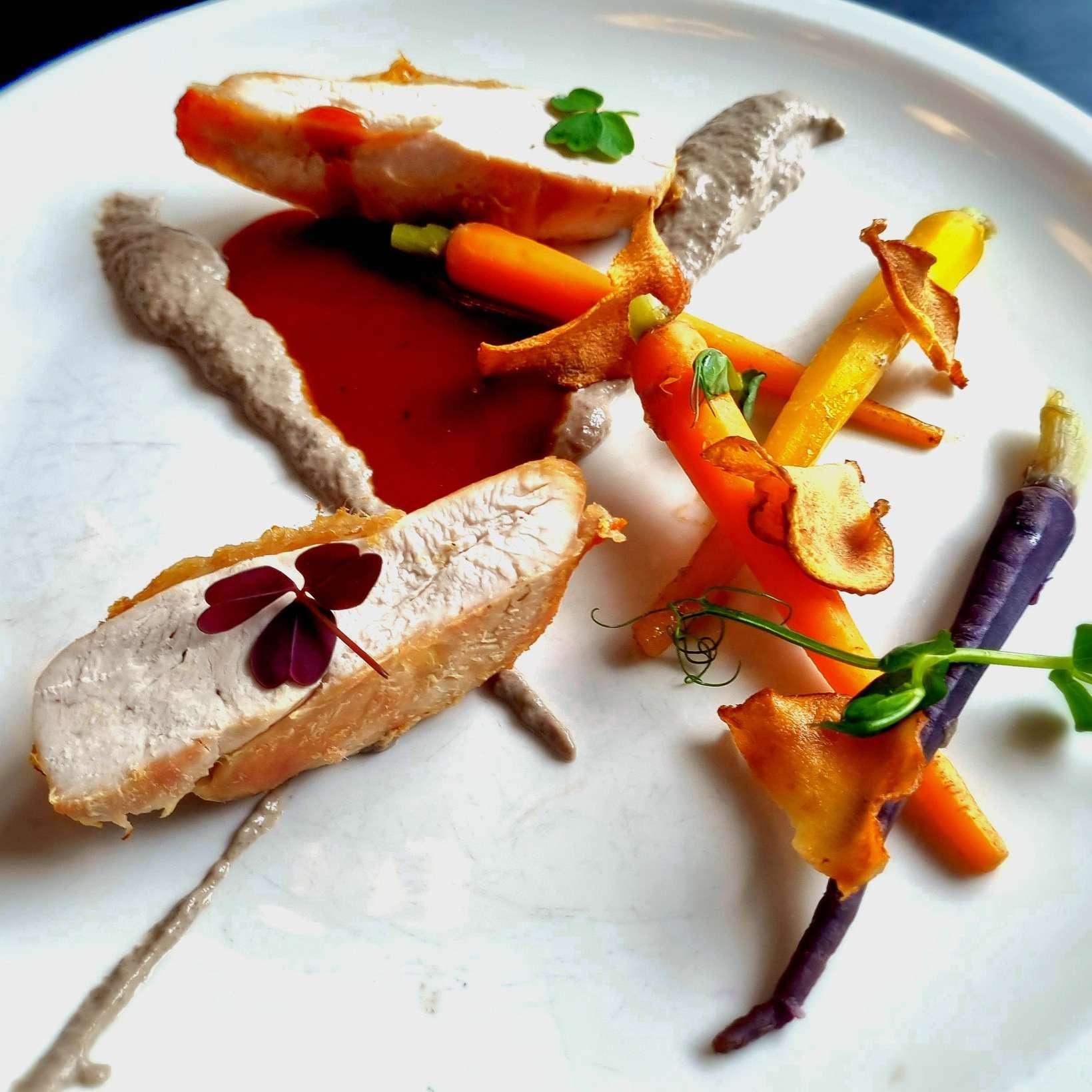 Two slices of cooked chicken breast garnished with small edible leaves, served with baby carrots, purple carrots, carrot crisps, a red sauce, and dollops of a grayish-beige puree on a white plate. The presentation is artistic and refined.