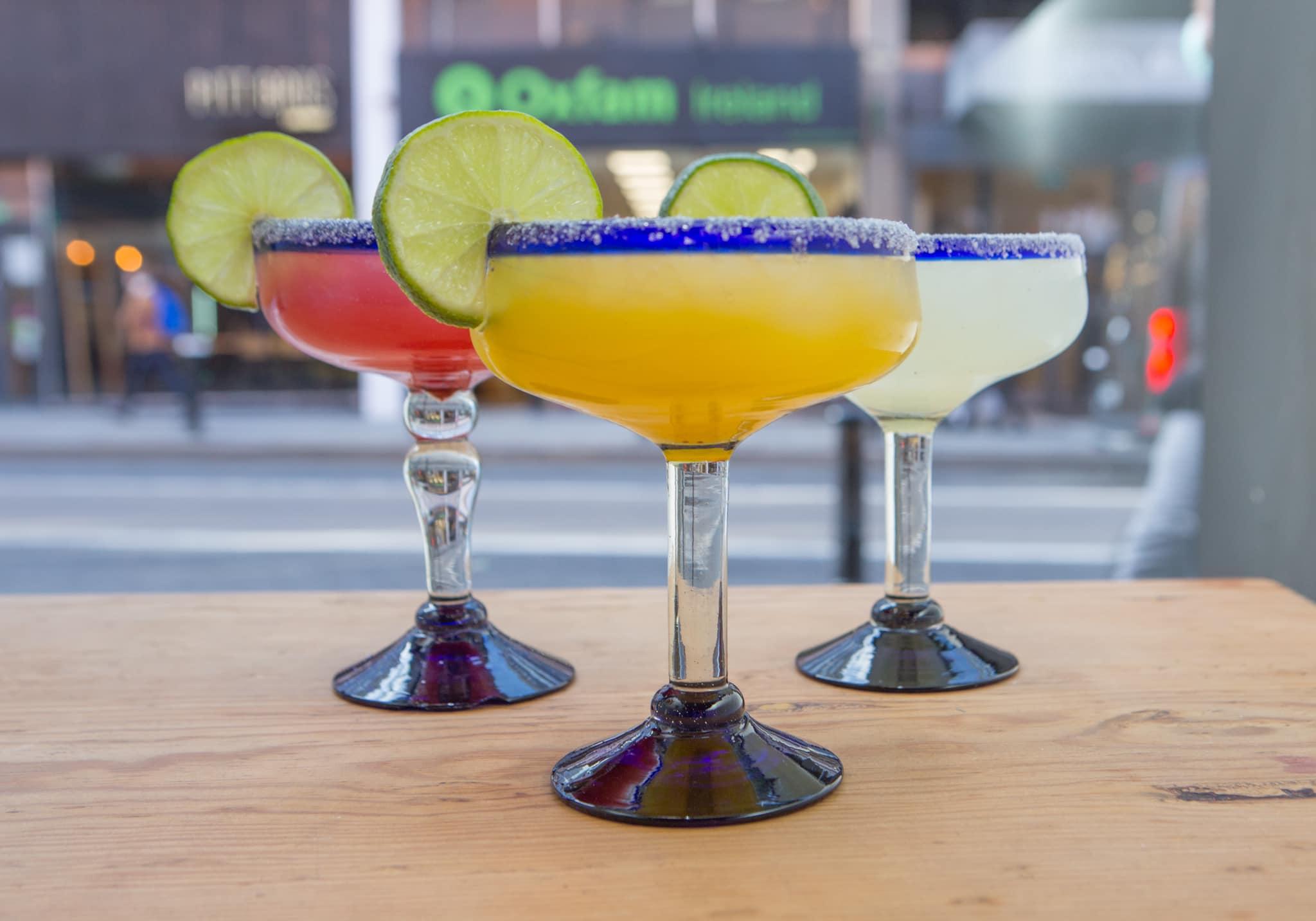 Three margarita glasses with salted rims and lime wedges, filled with red, yellow, and clear cocktails, sitting on a wooden table with a blurred city street background.