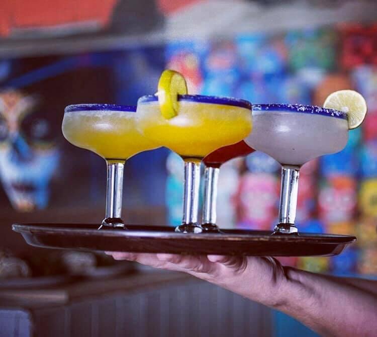 A hand is holding a tray with three margarita glasses. Two of the drinks are yellow with a salted blue rim and a lime wedge garnish, while the third drink is clear with a similar salted blue rim and a lime wheel garnish. The background is colorful and blurred.