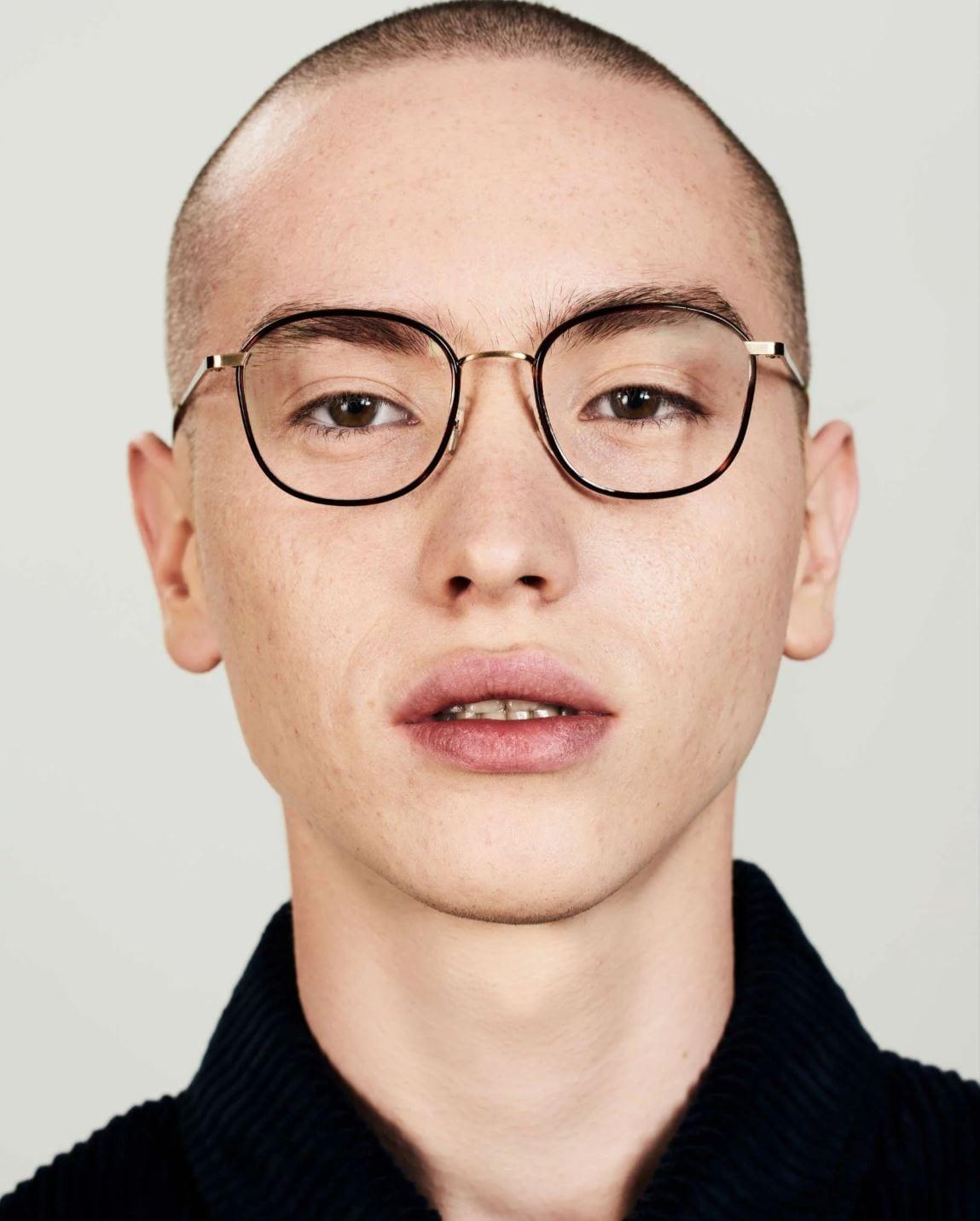 Close-up portrait of a person with a shaved head, wearing large round glasses with thin black frames and a dark sweater, looking directly at the camera with a neutral expression.