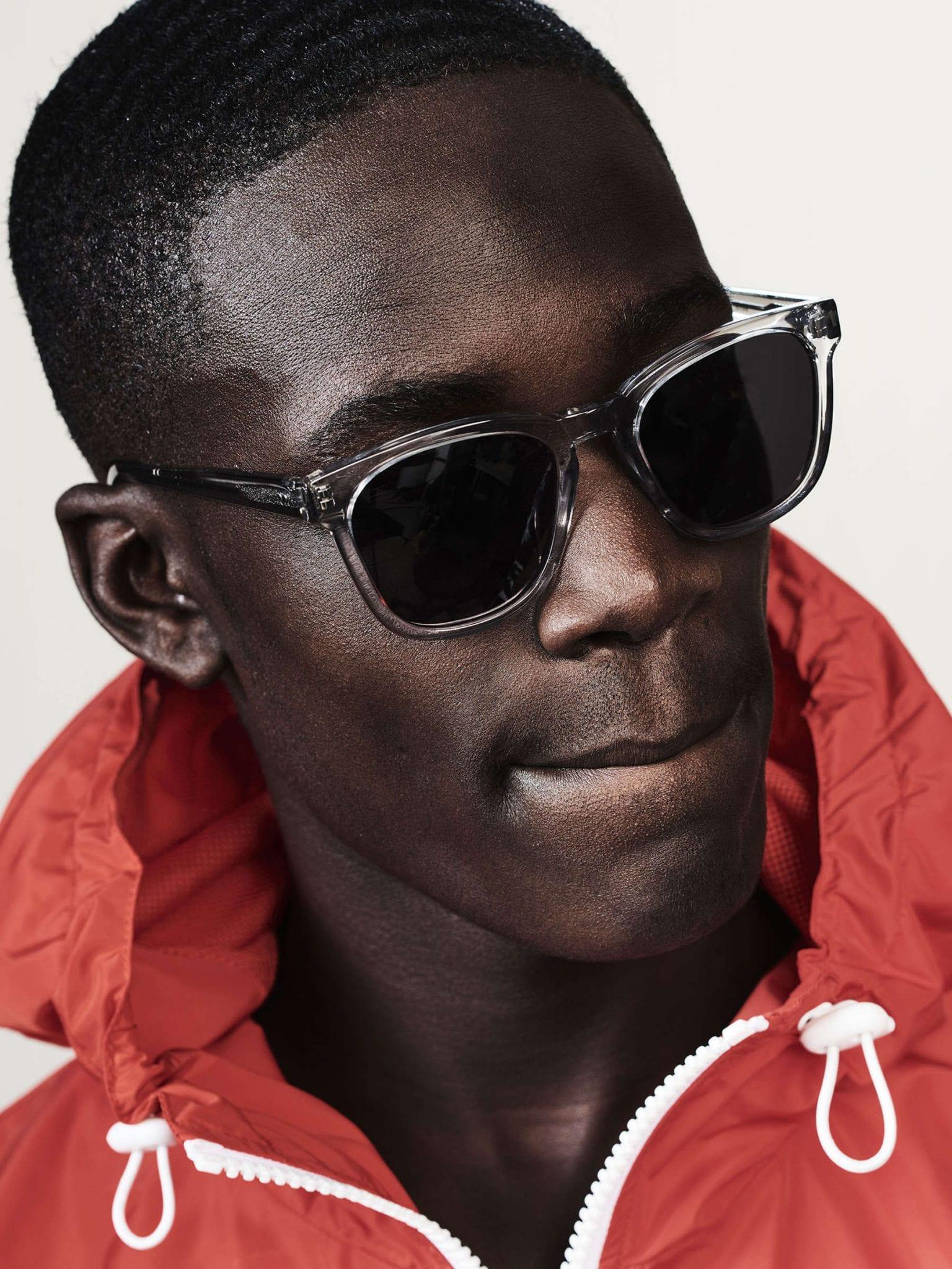 Close-up of a person wearing dark rectangular sunglasses with clear frames and a red zip-up jacket with a hood, looking slightly down and to the side with a neutral expression.