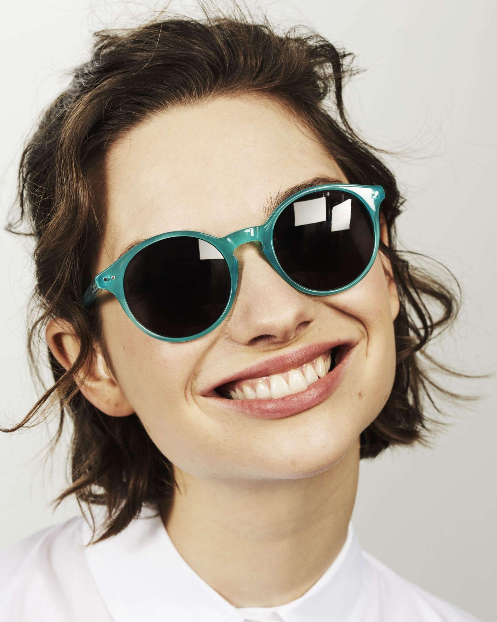 Close-up of a smiling person wearing round teal sunglasses with dark lenses and a white collared shirt, facing slightly to the side