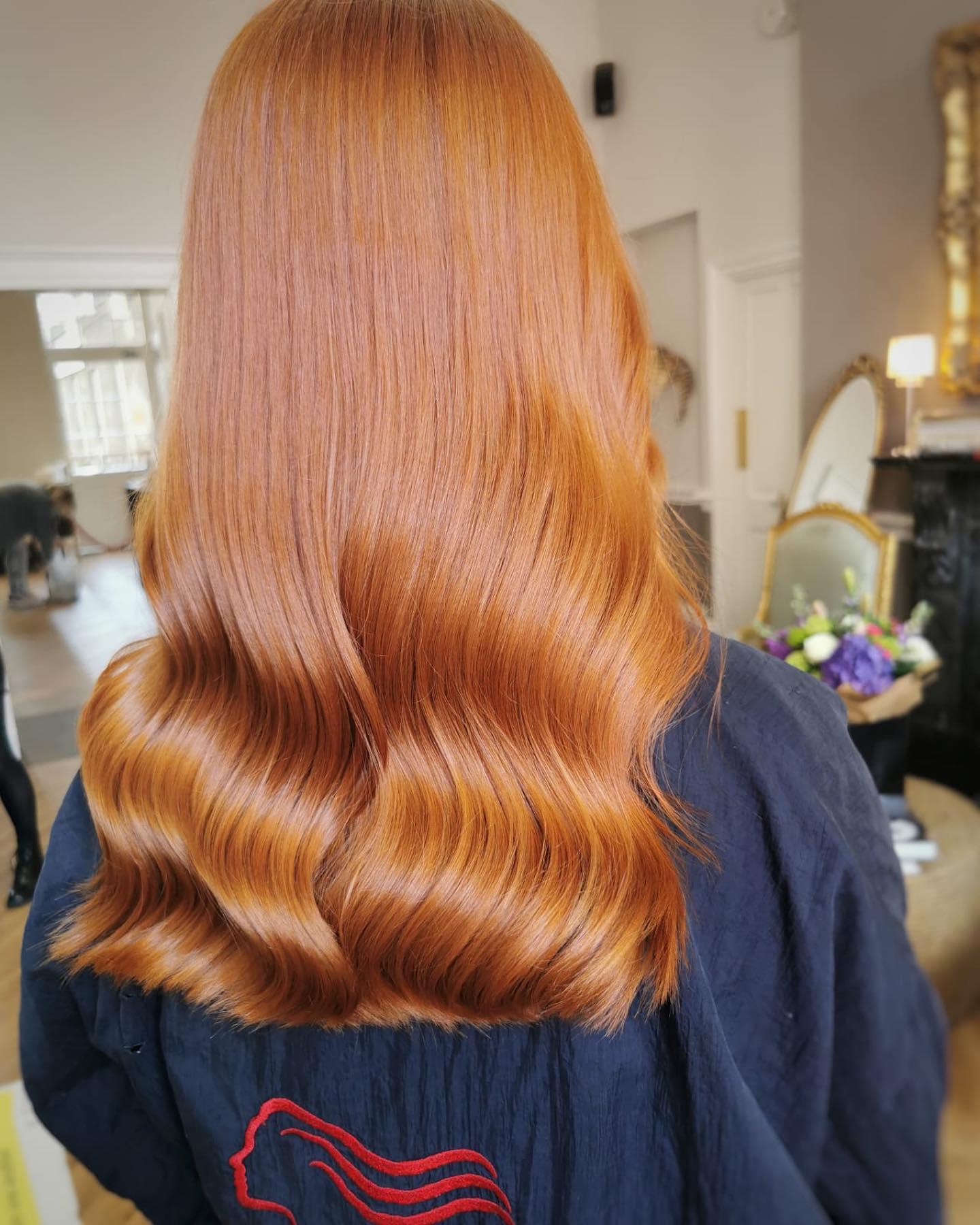 Back view of a woman with long, glossy copper-red hair styled in smooth, soft Hollywood waves. The hair is shiny and evenly colored, with a rich, vibrant finish. She is wearing a dark navy salon cape embroidered with a red line-art hair motif. The setting appears to be an upscale salon interior with a mirror, flowers, and warm lighting.