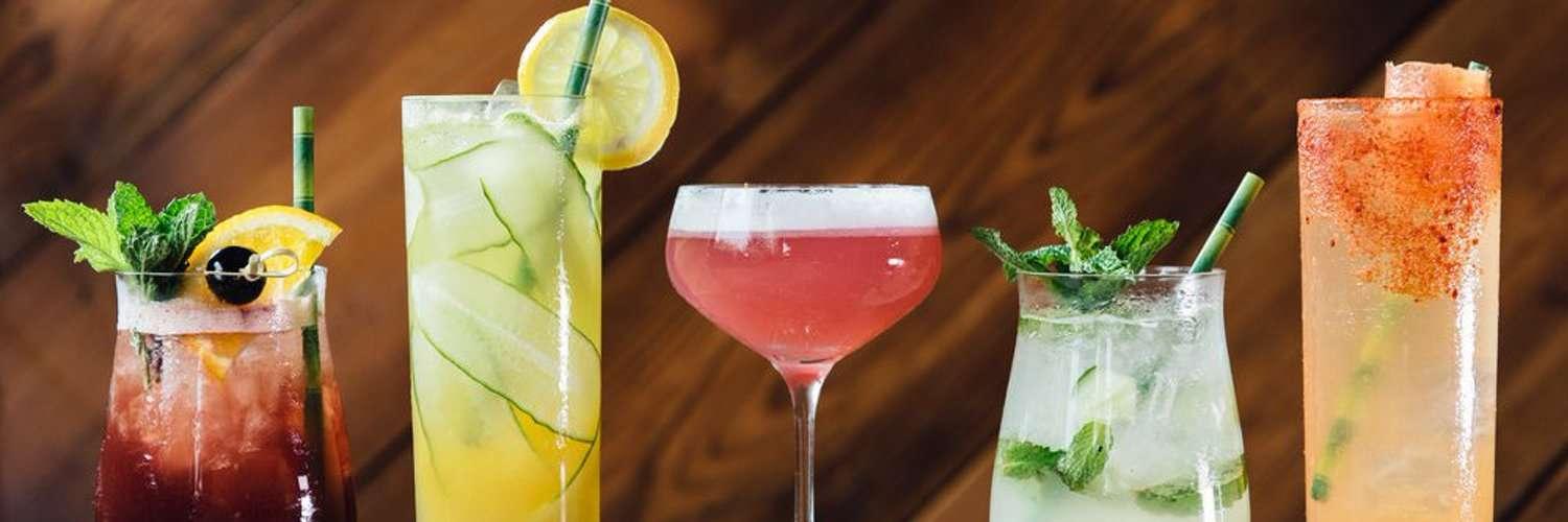 A row of five colorful cocktails displayed against a wooden background. From left to right: a dark red drink garnished with mint, orange slice, and cherry; a yellow cocktail with cucumber slices and a lemon wheel; a pink drink in a coupe glass with a foamy top; a mojito-style drink with mint and lime; and a tall orange cocktail with a red chili powder rim and a grapefruit wedge.