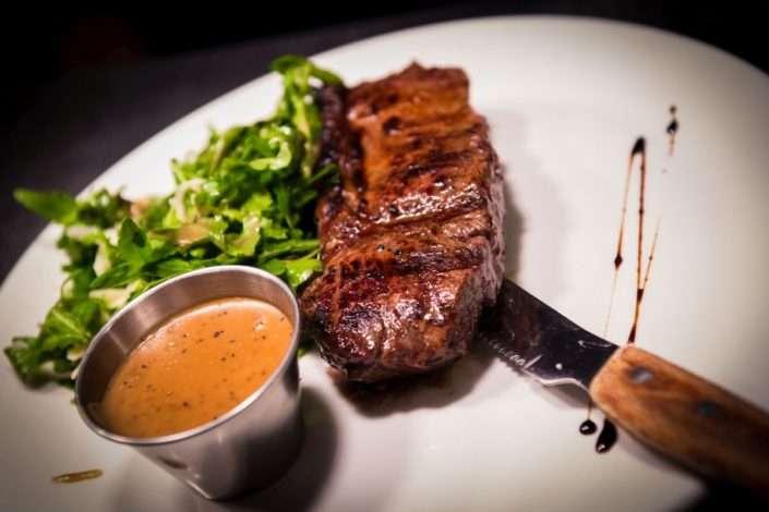 Grilled steak served on a white plate with a side of fresh green salad, a metal cup of peppercorn sauce, and a steak knife. A drizzle of dark balsamic glaze decorates the plate.