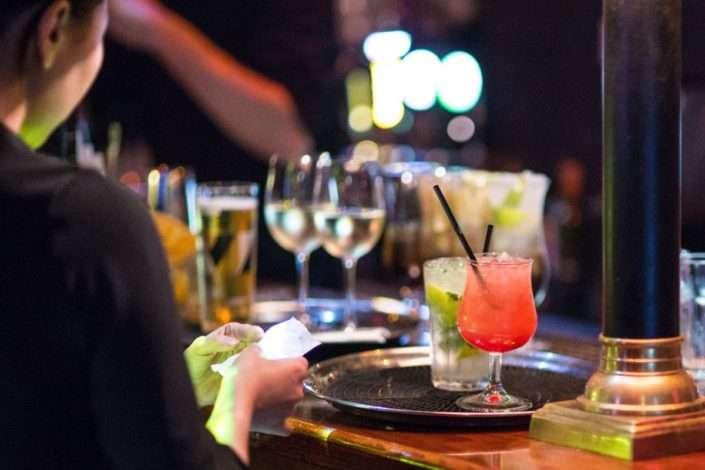 A server at a dimly lit bar prepares to deliver a tray of assorted drinks, including a red cocktail with a straw, a mojito with lime and mint, and glasses of white wine and beer in the background.