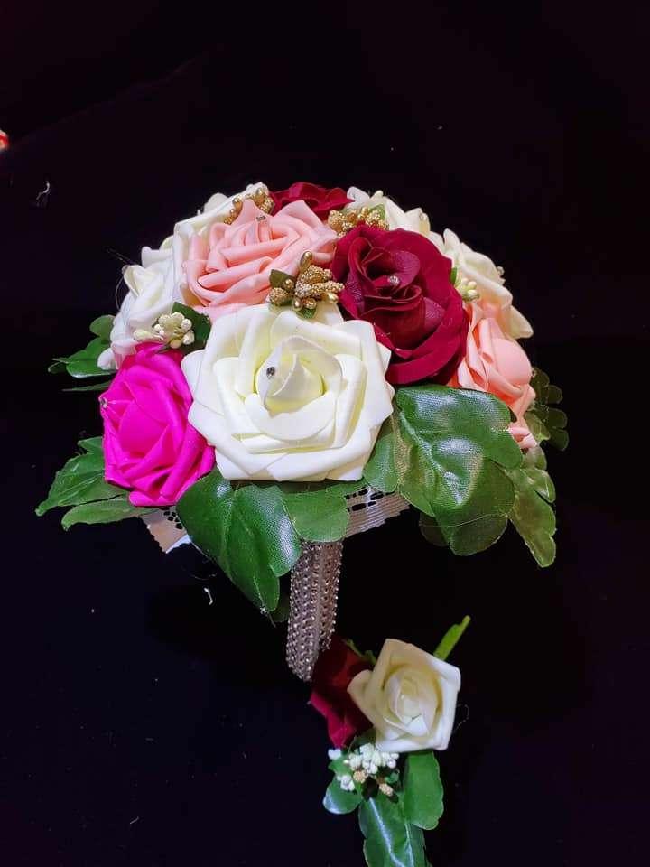 A vibrant artificial flower bouquet arranged with white, pink, peach, red, and magenta roses, accented with green leaves and small decorative buds, set against a black background. A matching boutonniere with red and white roses is placed below the bouquet.
