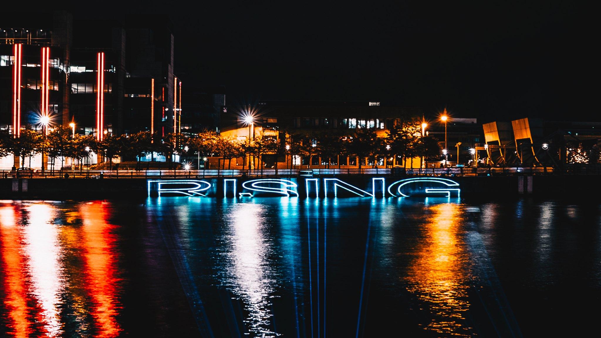 Neon light installation spelling the word “RISING” in blue, reflected on a river at night with illuminated office buildings and streetlights in the background.