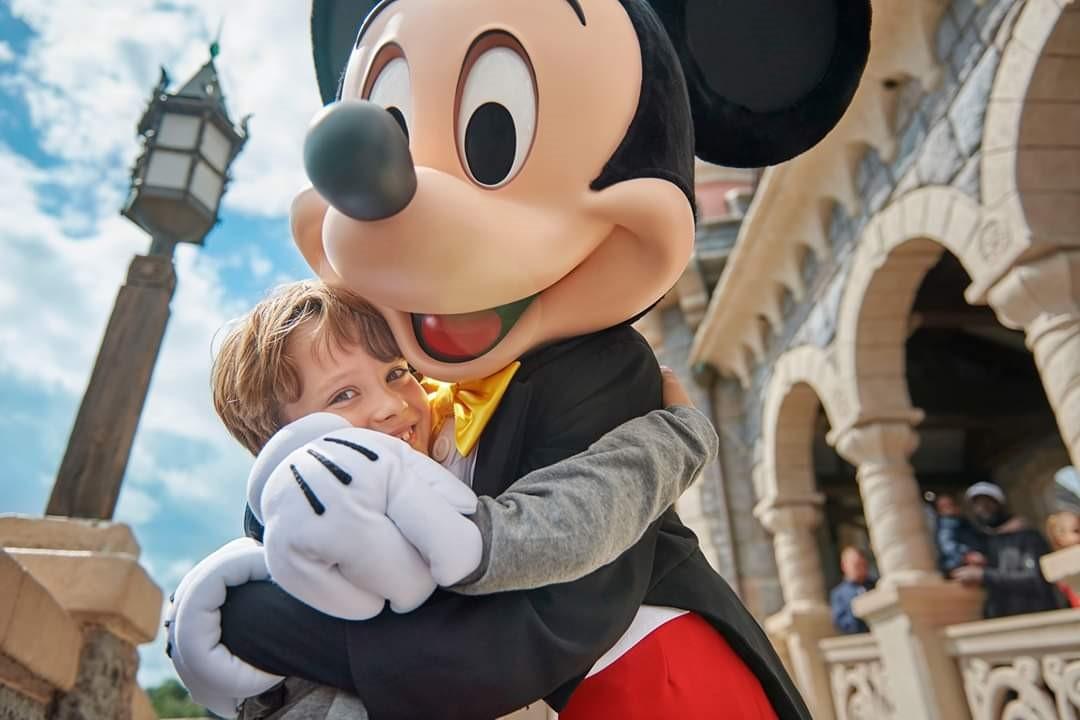 Young child hugging a person in a Mickey Mouse costume outside a castle-like building, with a lamppost and blue sky in the background.