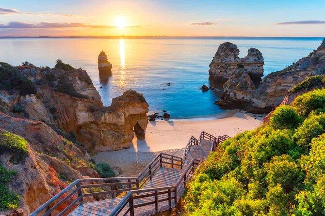 Sunset view of a sandy beach with unique rock formations and a wooden staircase leading down to the shore, surrounded by lush green bushes.