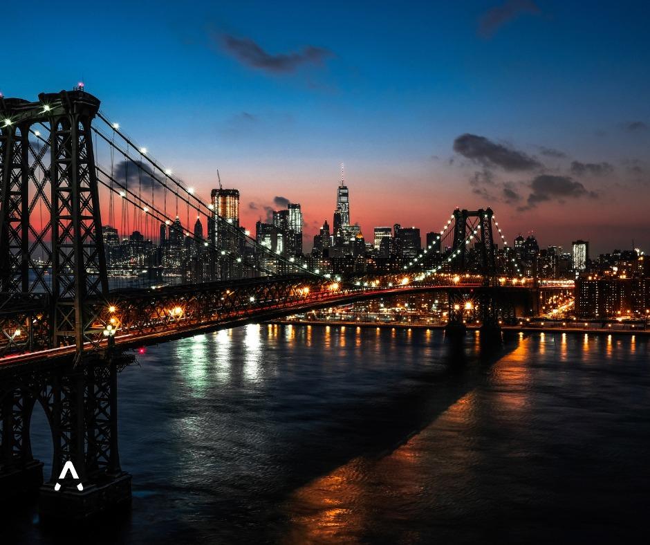 Evening view of the Manhattan skyline in New York City, with the illuminated Williamsburg Bridge spanning the East River under a colourful sunset sky.