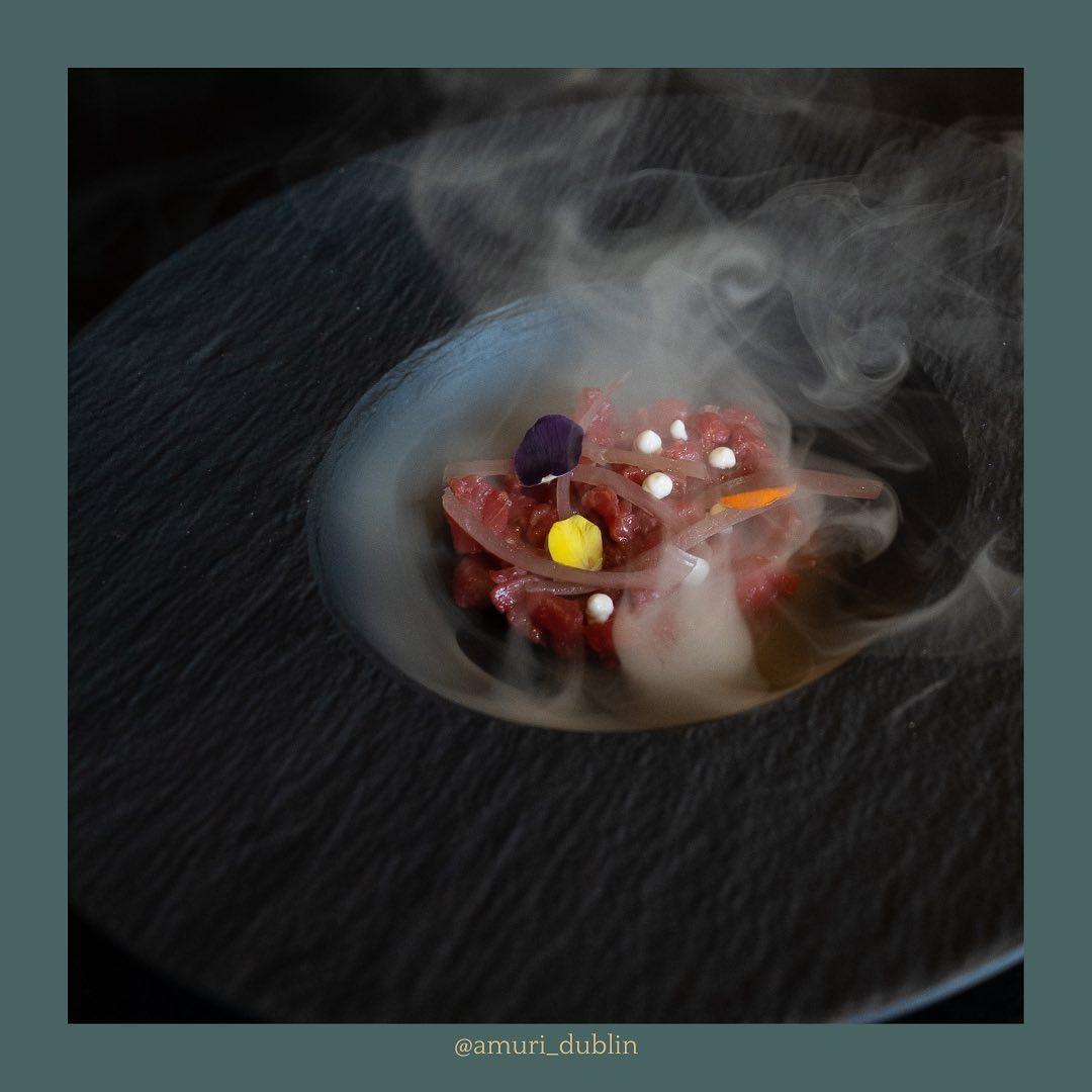 A gourmet dish served on a matte black plate, featuring a delicate portion of raw beef tartare garnished with thinly sliced onions, edible flowers, and white spheres, surrounded by swirling smoke for a dramatic presentation. Styled by @amuri_dublin.