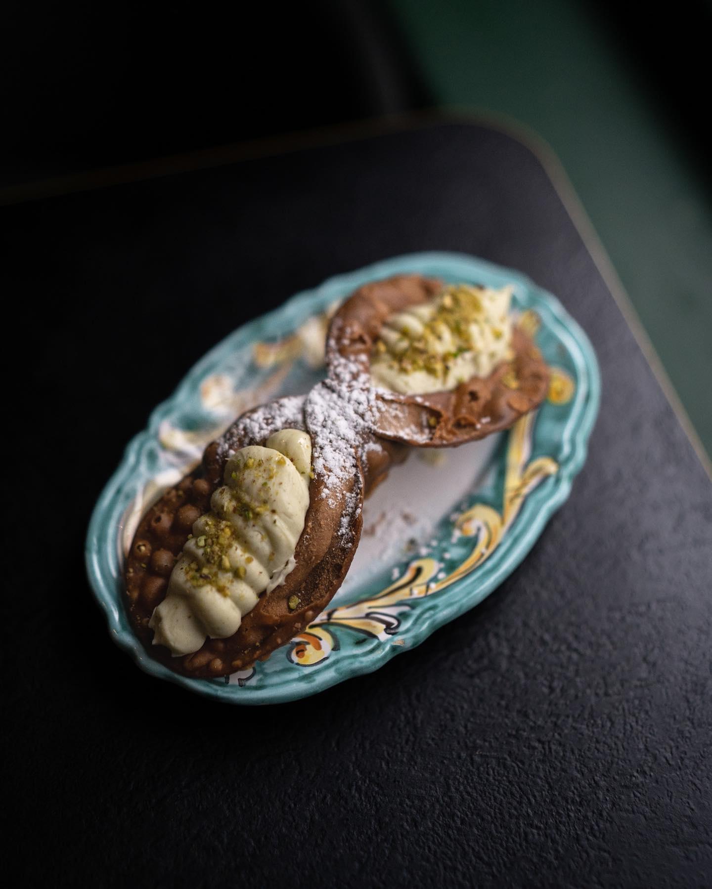 A pair of Sicilian cannoli filled with creamy ricotta, topped with crushed pistachios and powdered sugar, served on an ornate turquoise and white ceramic plate with yellow detailing, placed on a dark tabletop