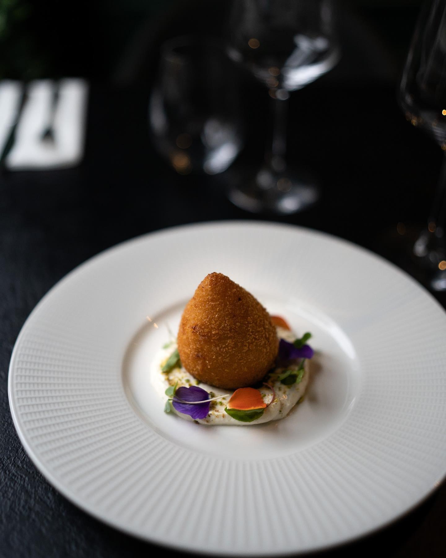 A single, golden-brown, pear-shaped arancini served on a white textured plate, resting on a bed of creamy sauce garnished with microgreens and edible flower petals, with a dark blurred dining setting in the background.