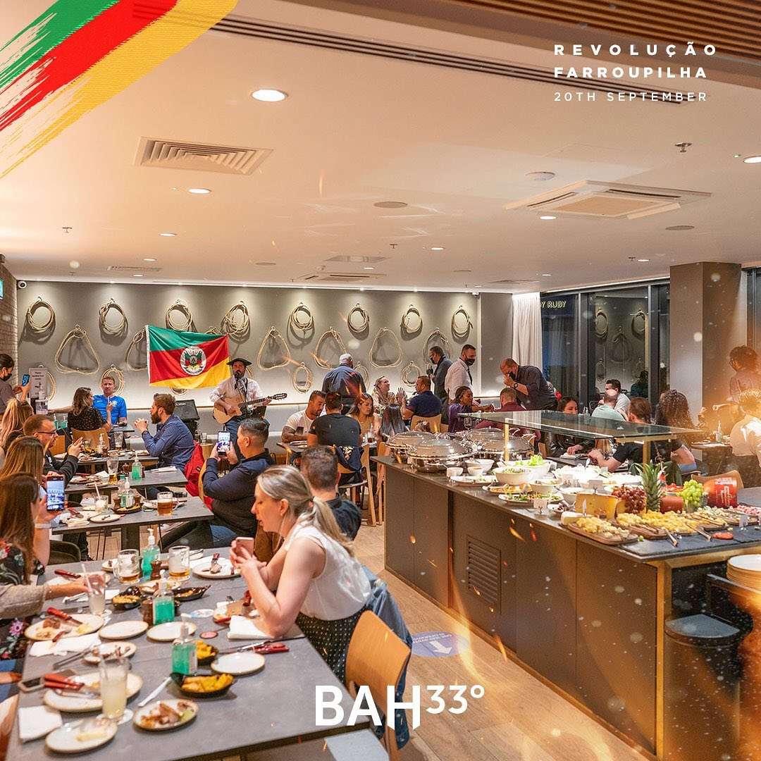 A lively indoor gathering where many people are seated at tables, enjoying food and drinks. In the background, a musician is playing guitar near a wall decorated with coiled ropes and a colorful flag. A large buffet table is filled with various dishes, fruits, and plates, creating a warm and inviting atmosphere. The room is well-lit, and the overall mood feels social and festive, with guests engaged in conversation and taking photos.