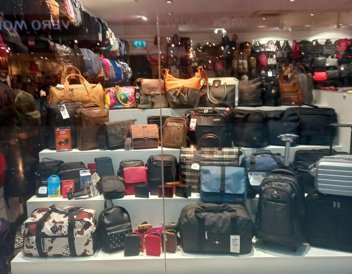 Storefront display of assorted bags, including leather handbags, travel duffels, backpacks, briefcases, and rolling suitcases, arranged on tiered shelves behind glass.