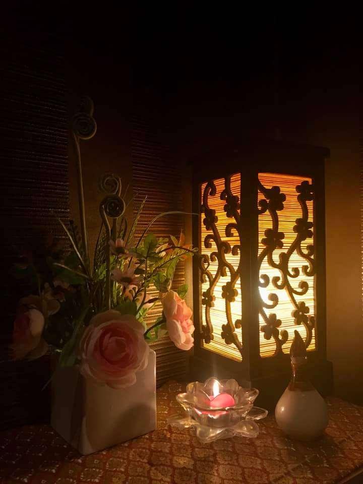 A warm-lit decorative lantern with floral cutouts, a small candle in a glass holder, a white vase with pink flowers, and a small bottle on a patterned surface.