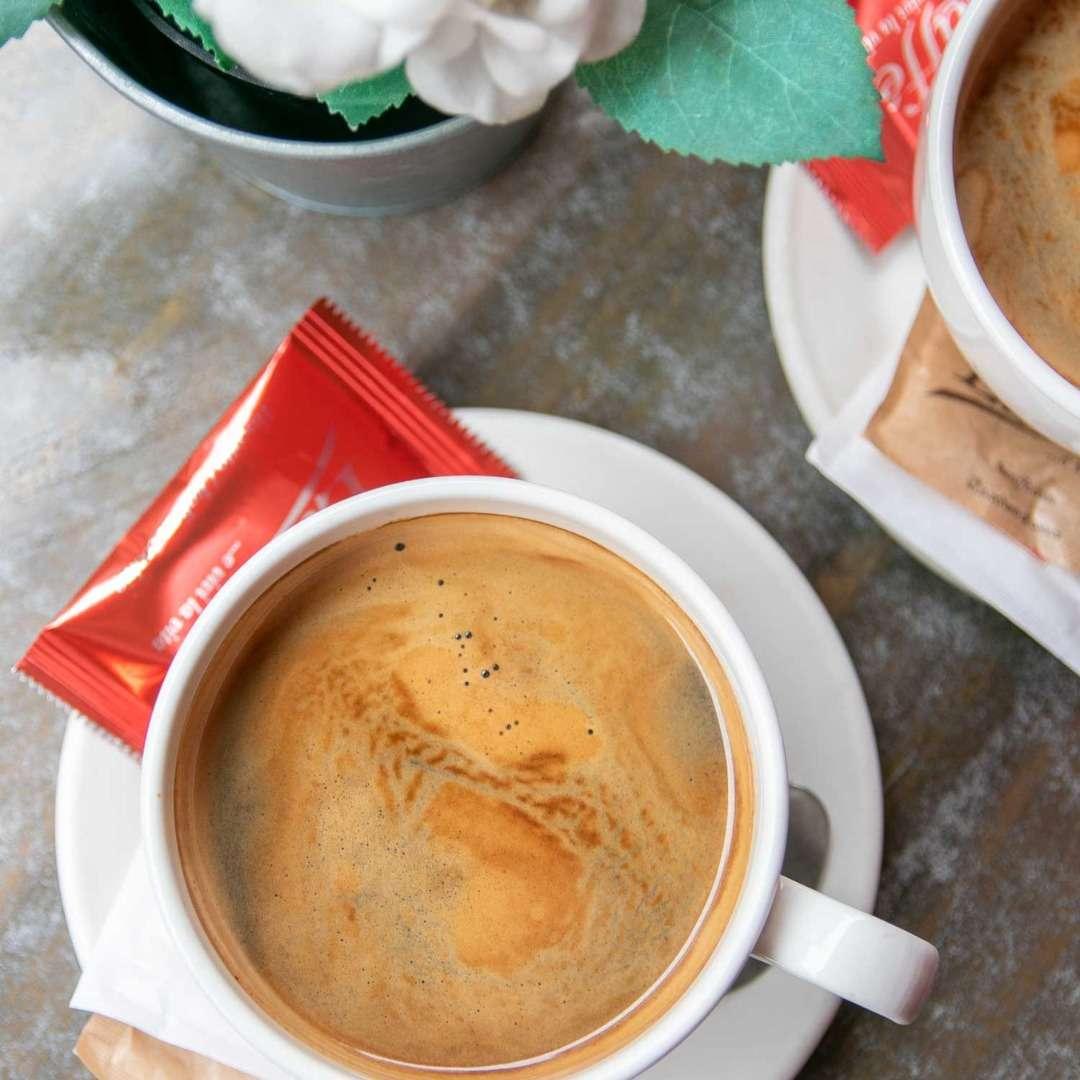 This image captures two cups of freshly brewed coffee, each served on a saucer with a small wrapped biscuit on the side. The rich crema swirls on top suggest a strong, aromatic brew. A decorative flower pot adds a touch of freshness to the cozy café setting.