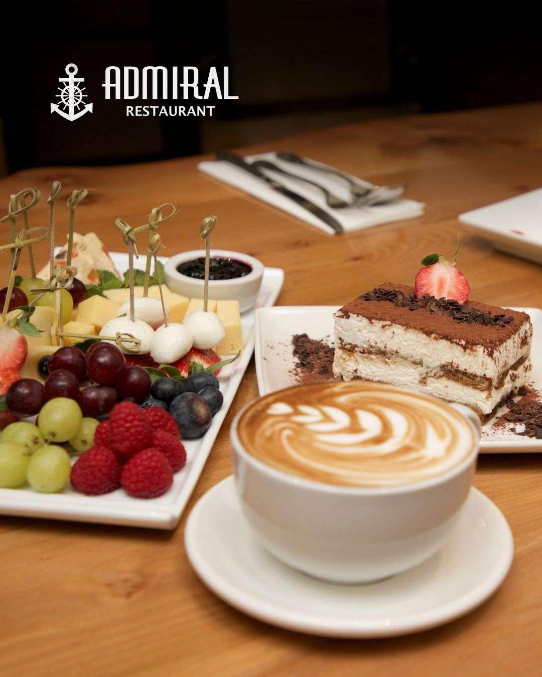 Assorted fruit and cheese platter with skewers, tiramisu topped with a strawberry, and a cappuccino with latte art, all served on white plates on a wooden table.