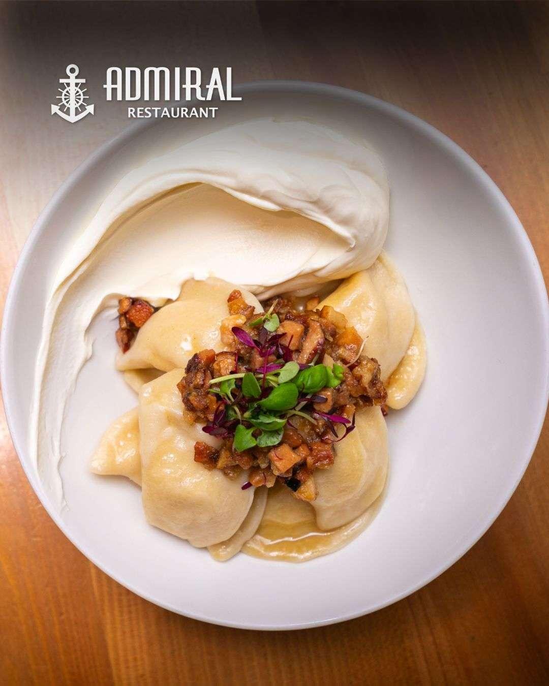 Bowl of handmade dumplings topped with crispy diced bacon and microgreens, served with a generous dollop of sour cream on the side.