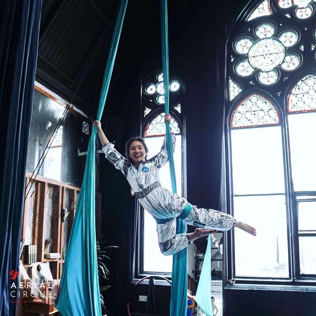 Smiling woman in a shiny silver costume performing an aerial pose using blue aerial silks in front of large stained glass windows inside a studio. The logo "Aerial Cirque Dublin, Ireland" appears in the bottom left corner.