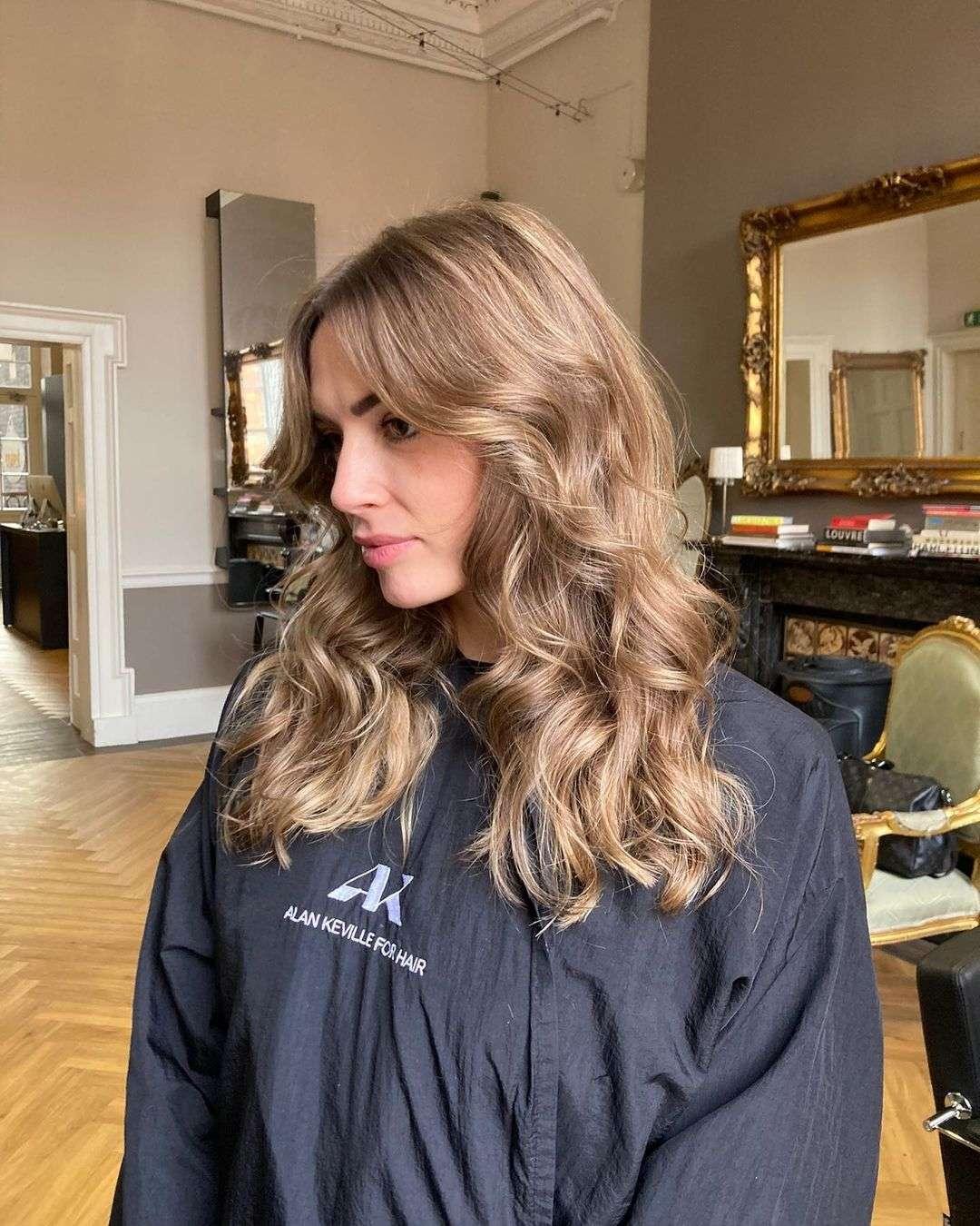 A woman with long, voluminous light brown hair styled in soft, loose waves and curtain bangs. She is wearing a black salon cape with the logo "AK Alan Keville for Hair" embroidered on it. The setting is an elegant hair salon with a herringbone wood floor, ornate gold-framed mirror, and neutral-toned walls.