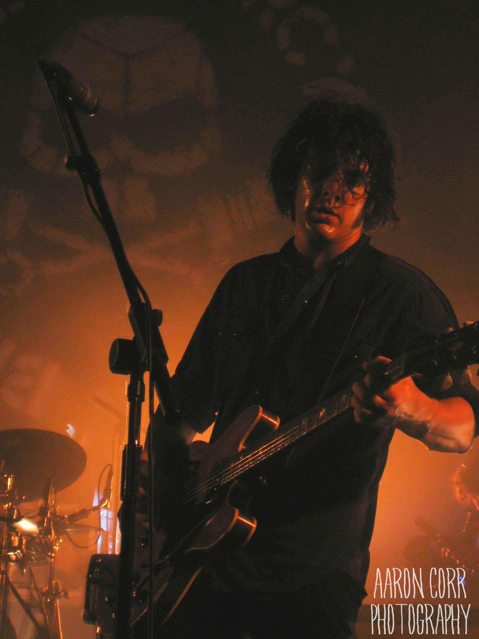 A musician playing an electric guitar on stage under warm orange lighting. The musician has dark, curly hair partially covering their face and is wearing a dark shirt. In the background, a drum set is visible. The photo has a watermark in the bottom right corner that says ‘Aaron Corr Photography.