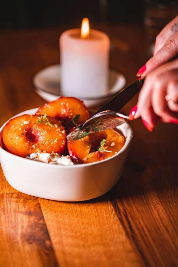 A hand holding a spoon over a dessert bowl filled with roasted peach halves on a bed of soft cheese or cream, with a lit candle in the background.