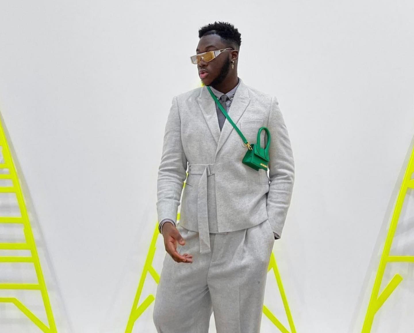 Stylish man posing against a white backdrop with neon yellow ladder-like structures. He is wearing a light gray tailored suit with a belted jacket, a gray shirt underneath, gold-framed sunglasses, and a small bright green crossbody bag. One hand is in his pocket while the other gestures casually
