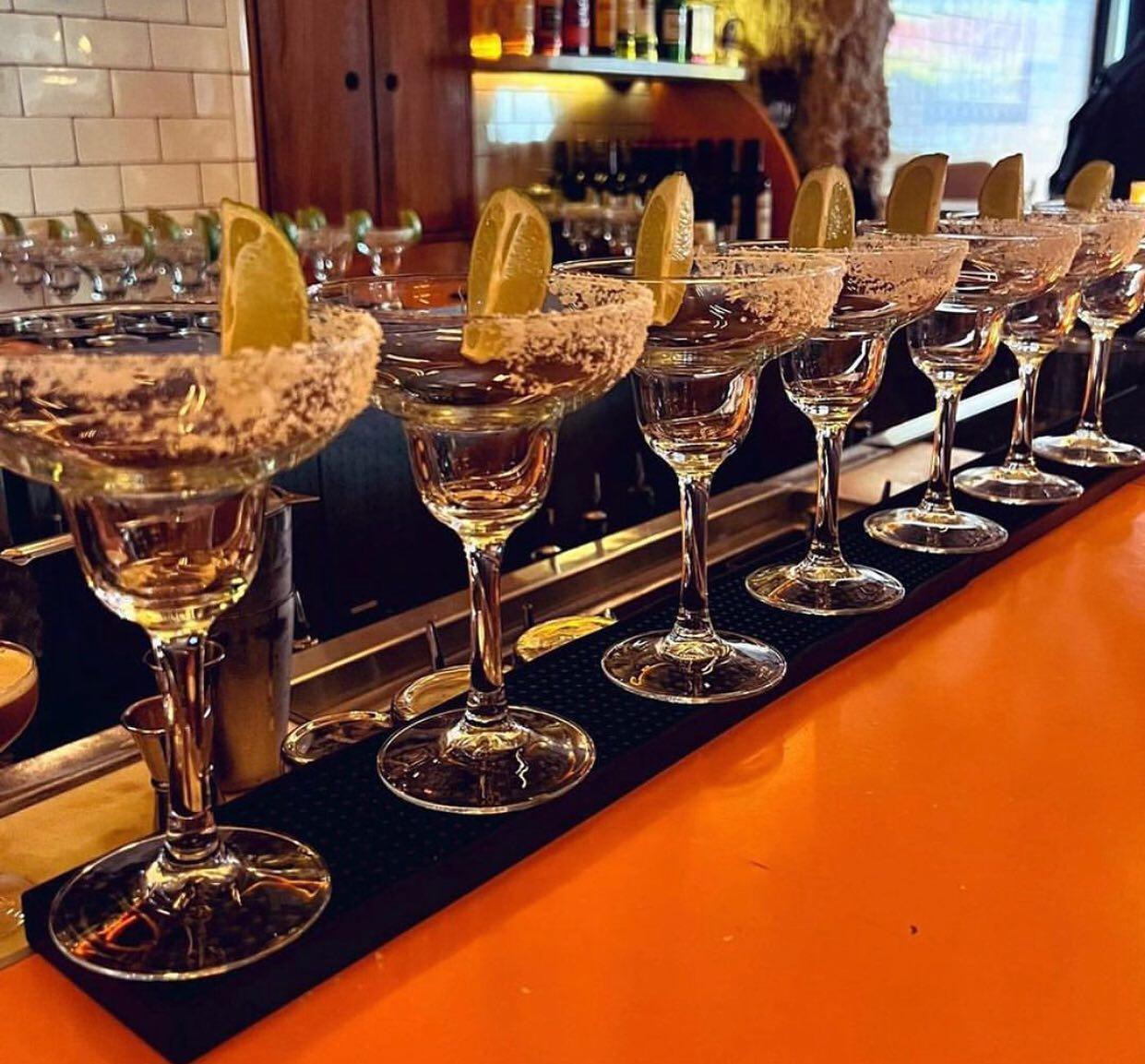 A row of margarita glasses lined up on a bar counter, each rimmed with salt and garnished with a wedge of lime. The bar in the background has bottles and glassware displayed on shelves.