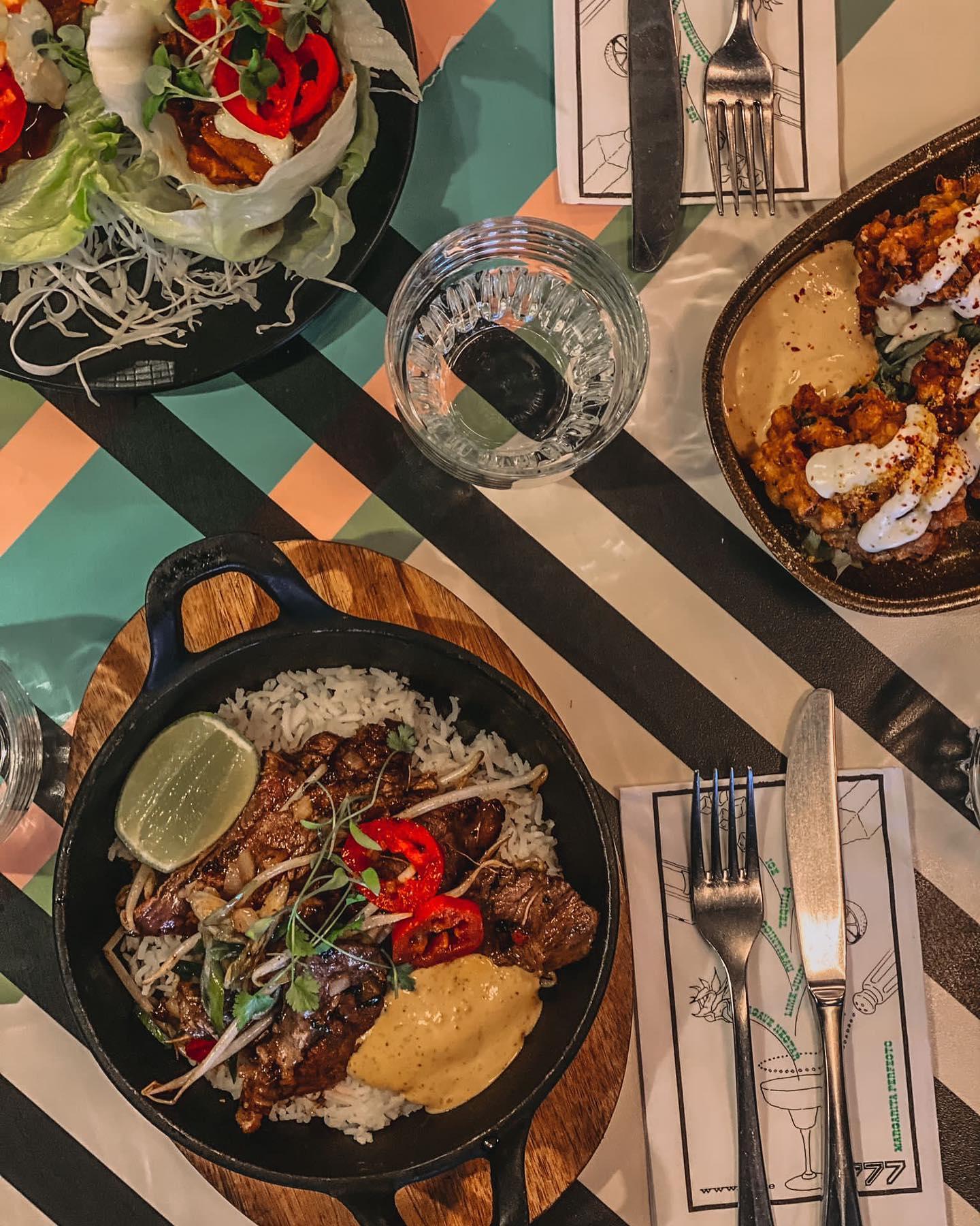 An overhead view of a colorful table set with three modern Mexican dishes. At the bottom center, a cast iron skillet on a wooden board contains grilled beef slices over white rice, topped with red chili slices, microgreens, a dollop of creamy yellow sauce, and a lime wedge. To the top left, lettuce cups filled with shredded vegetables and spicy toppings are arranged on a black plate. At the top right, a brown dish holds crispy cauliflower bites drizzled with white sauce and served with a beige dip.