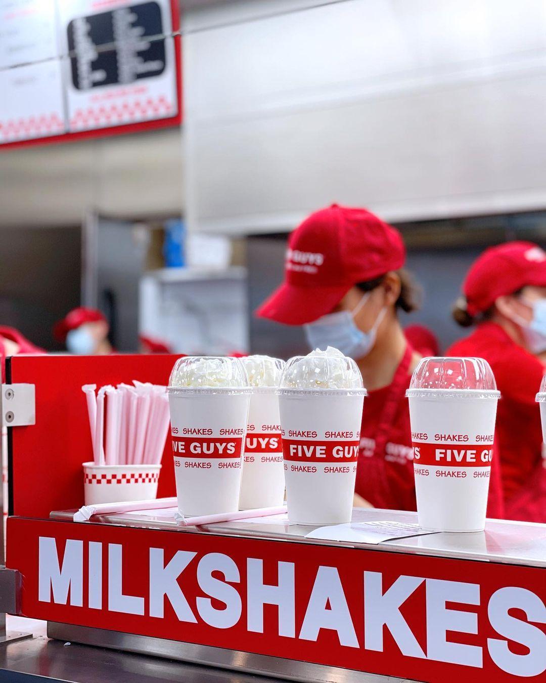 A row of creamy milkshakes topped with whipped cream sits ready on a red counter labeled "MILKSHAKES" at Five Guys. Each shake is in a branded white cup with the red "FIVE GUYS" and "SHAKES" text repeated around the middle. In the background, employees in red uniforms and caps work behind the counter, some wearing face masks, highlighting the busy and clean environment of the Five Guys kitchen.