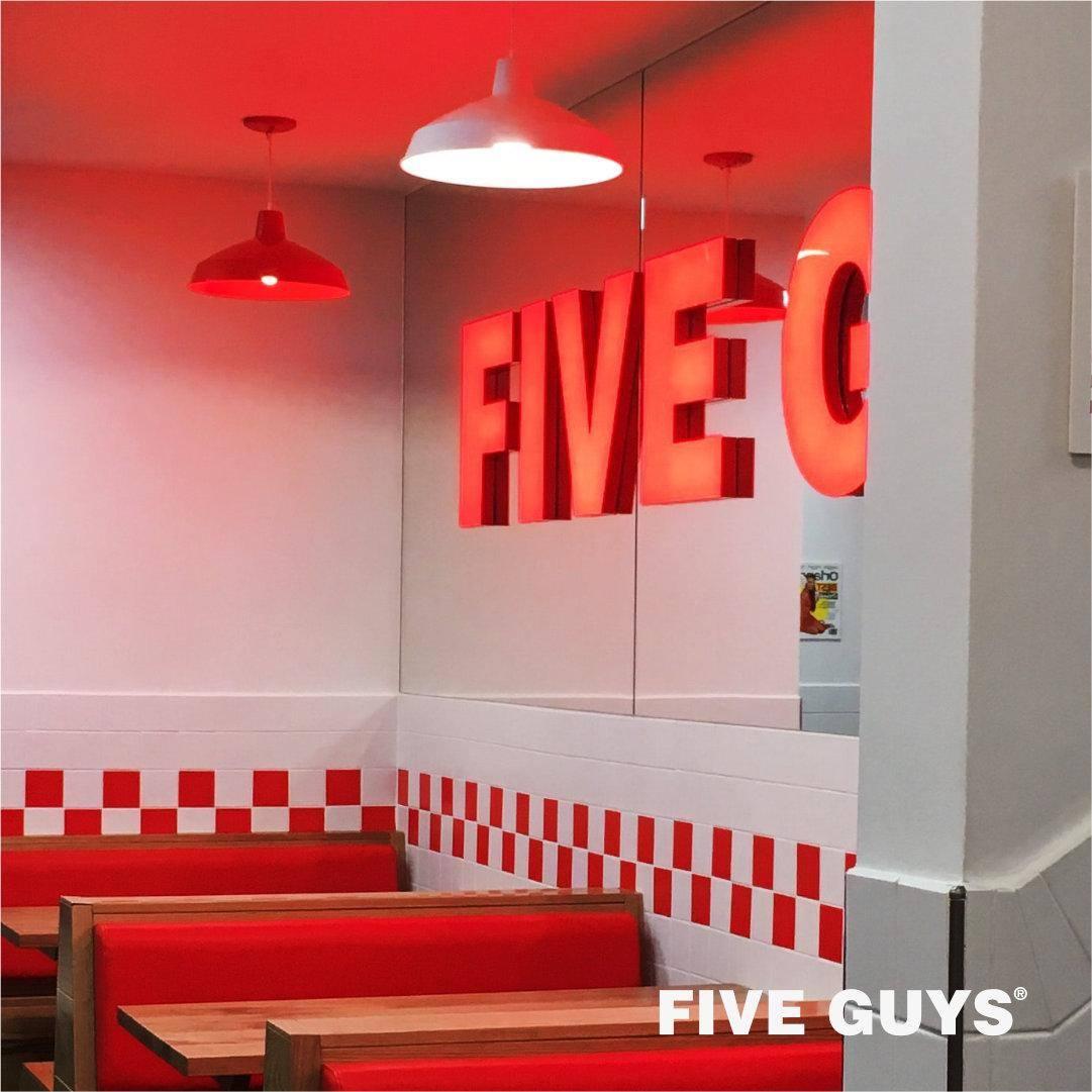 Interior of a Five Guys restaurant featuring red cushioned booths, white tiled walls with a red checkerboard pattern, and red and white pendant lights. A large illuminated "FIVE GUYS" sign is reflected in a wall mirror.