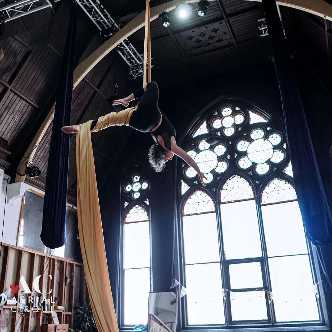 Aerial performer suspended upside down in a dramatic split pose using golden aerial silks, framed by tall stained glass windows and high ceilings inside a studio. The "Aerial Cirque Dublin, Ireland" logo is visible in the bottom left corner.