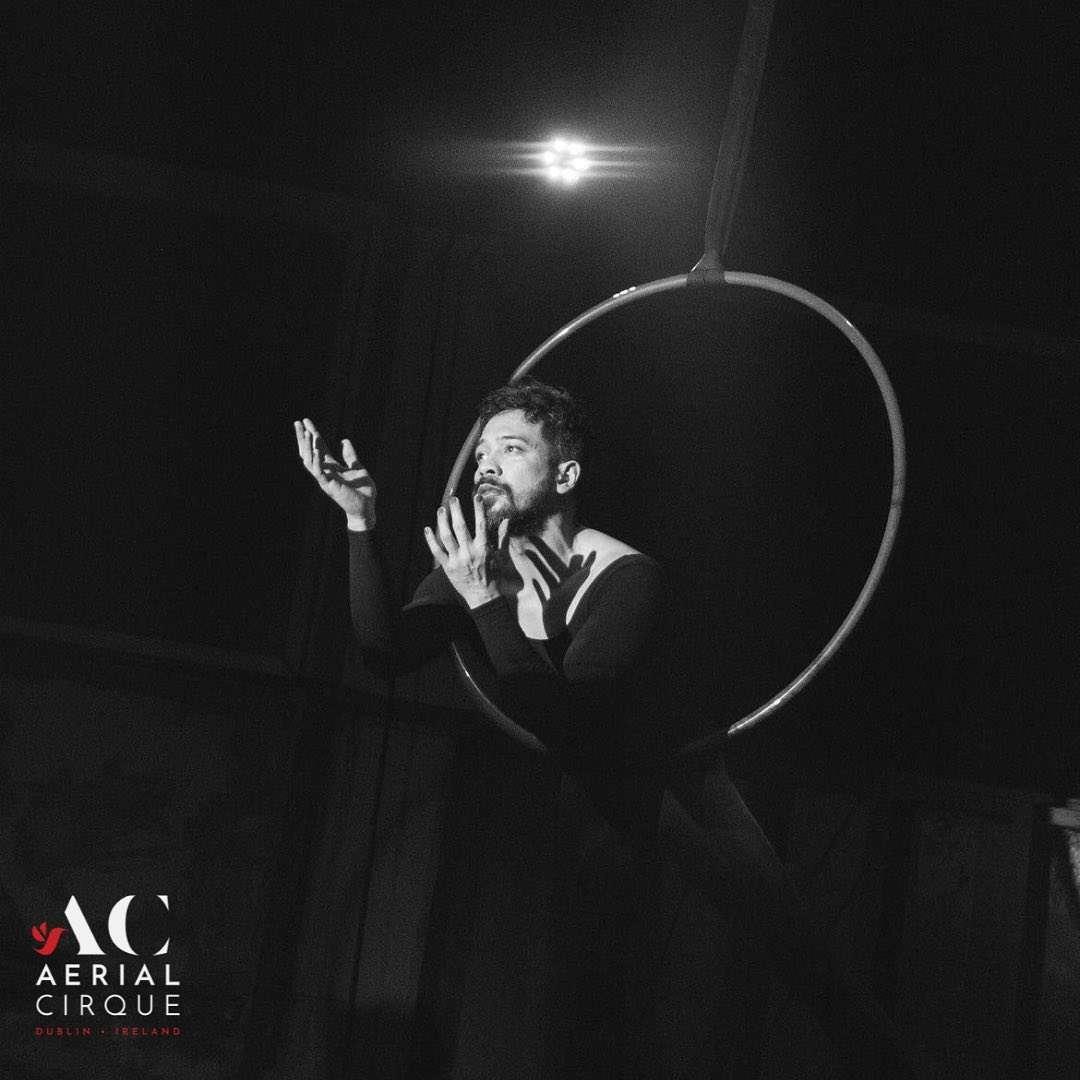 Black and white photo of a male aerial performer in a dramatic pose inside a suspended aerial hoop (lyra), with expressive hand gestures and a focused facial expression under a spotlight. The "Aerial Cirque Dublin, Ireland" logo appears in the bottom left corner.